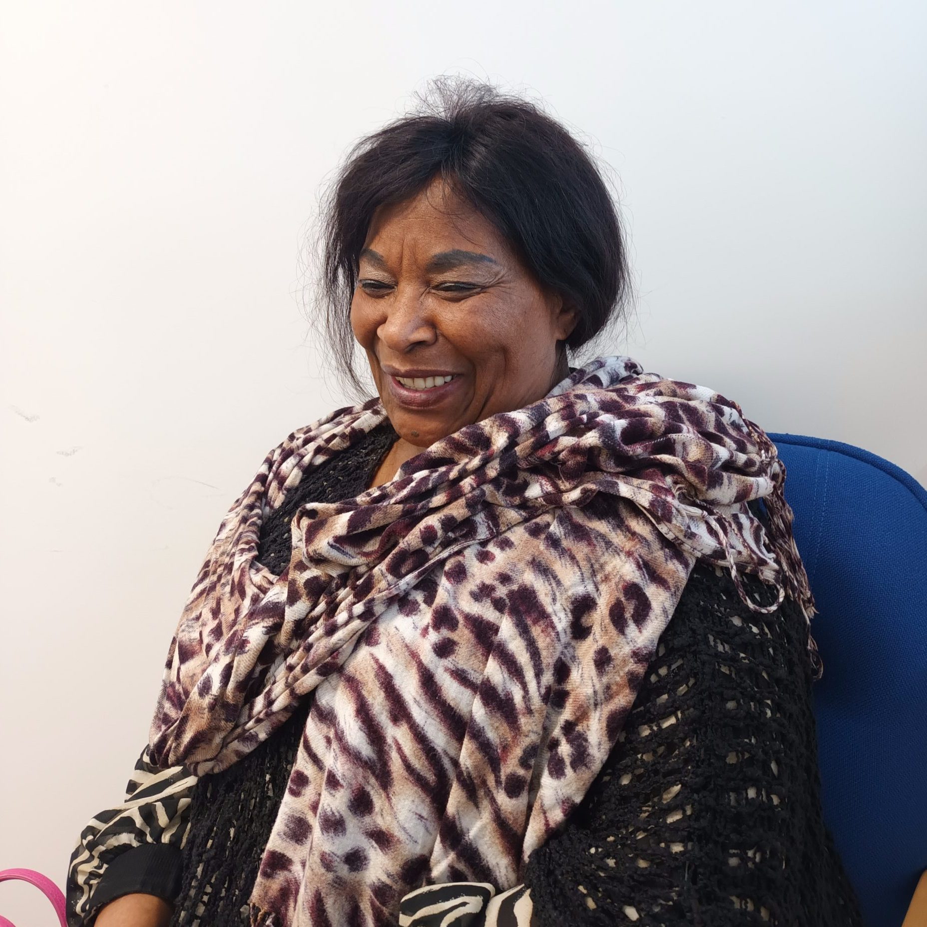 Volunteer mary is sat at an office desk with a big smile. She has black hair which is tied back and is wearing a black long sleeved top with a purple and white shawl wrapped around her.