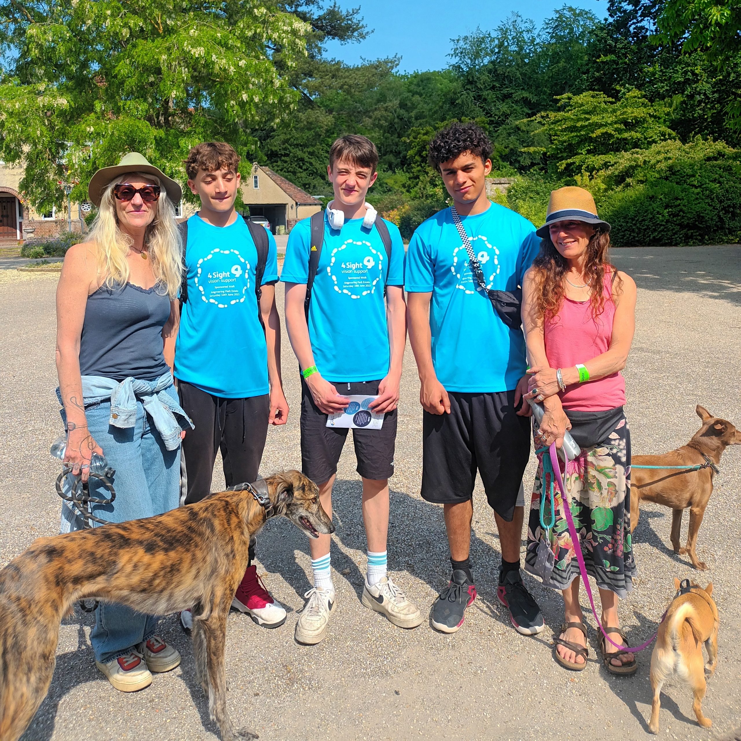 A group of walkers (2 women and 3 teenage boys) are stood in a line together with 3 dogs smiling towards the camera before they set off on their walk.