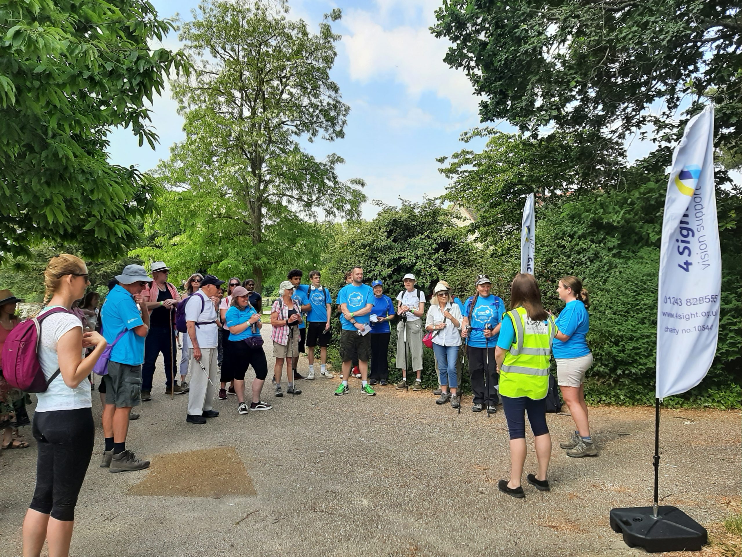 a large group of walkers in their gear are stood around listening to the walk briefing under the warm blue skies.